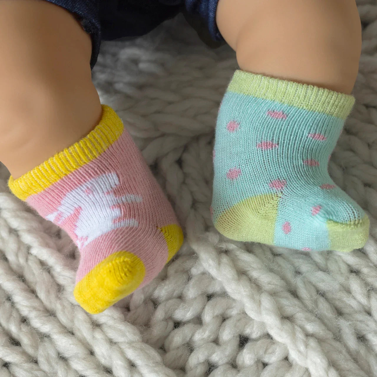 To babydukke føtter med fargerike sokker, rosa med motiv og blå med prikker, på strikket teppe